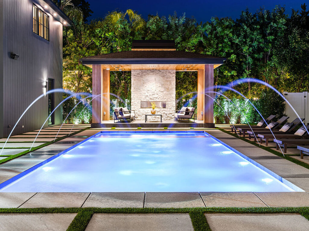 A swimming pool with illuminated water and fountain features in a landscaped area at night.